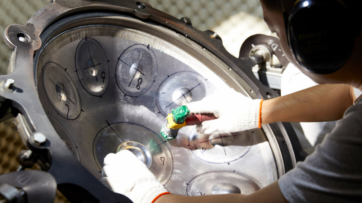 Hände in Arbeitshandschuhen bearbeiten eine angezeichnete Handpan-Schale in einer Spannvorrichtung