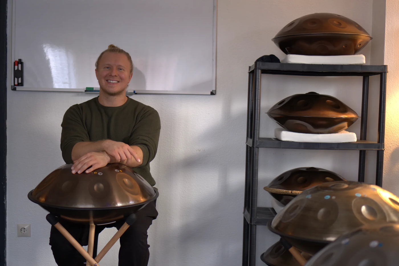 Handpan-Kursleiter sitzt im Studio an eine Handpan gelehnt, im Regal weitere Handpans